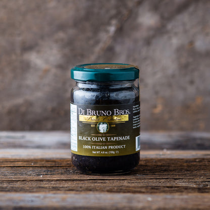 A jar of Di Bruno Bros. Black Olive Tapenade placed on a wooden surface with a neutral background.
