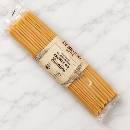 A package of Di Bruno Irons Bronze Die No. 10000 Italian Pasta, featuring light brown pasta strands inside a clear plastic wrapper with a beige paper label, resting on a white surface.