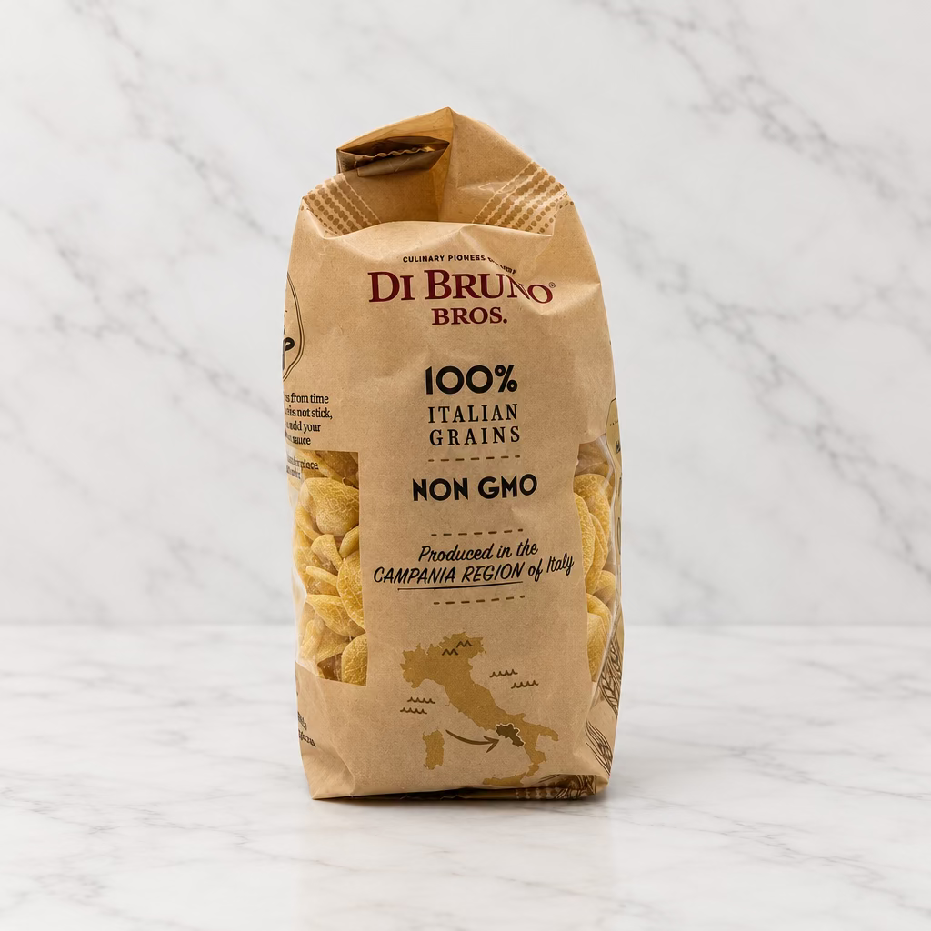 A bag of Di Bruges Bros. 100% Italian Grains Non Grooved pasta, standing upright on a white surface against a white background with a spotlight overhead.