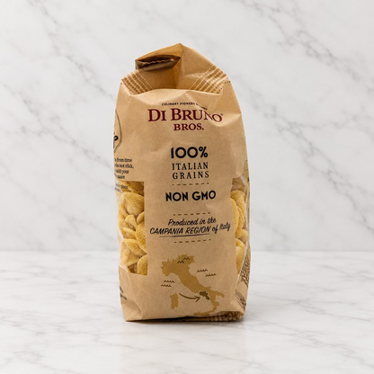 A bag of Di Bruges Bros. 100% Italian Grains Non Grooved pasta, standing upright on a white surface against a white background with a spotlight overhead.