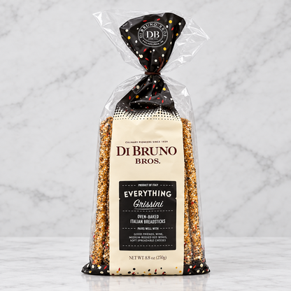 A bag of Di Bruno Bros. Everything Crissini oven-baked Italian breadsticks, shown in a clear plastic package with a cream and dark brown label and tied dark brown top, on a transparent checkered background.