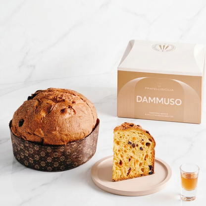 A loaf of Dammuso panettone bread in a dark patterned paper liner next to a slice on a plate and a small glass, with the branded box behind on a light tabletop.
