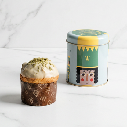 A decorated panettone in a patterned wrapper sits beside a cylindrical tin canister with a colorful nutcracker illustration and green/yellow label, set on a beige surface with a warm brown backdrop.