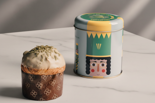 A decorated panettone in a patterned wrapper sits beside a cylindrical tin canister with a colorful nutcracker illustration and green/yellow label, set on a beige surface with a warm brown backdrop.