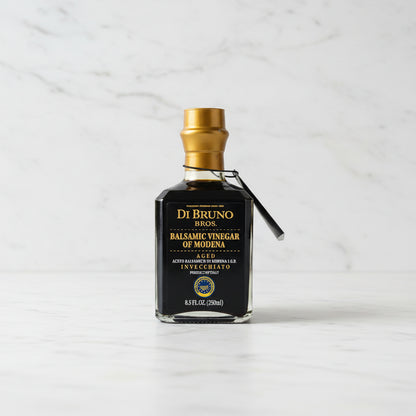 Bottle of Di Bruno Bros Balsamic Vinegar of Modena on a gray surface with a white tiled wall in the background.