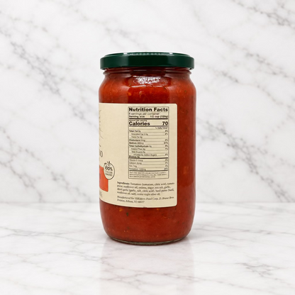 A glass jar of pasta sauce with a dark green lid and a beige nutrition facts label, shown from the back on a white studio background.