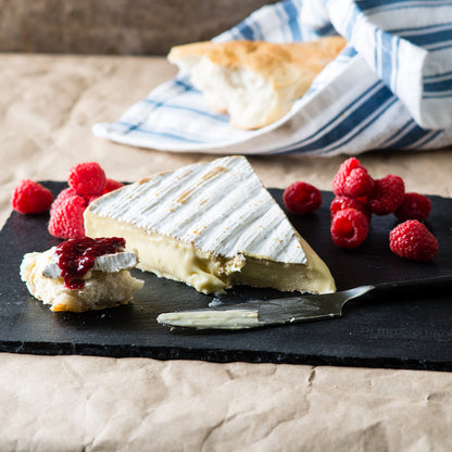 A wedge of brie cheese with a rind, garnished with jam and accompanied by fresh raspberries, crostini, and a side of bread on a black slate platter.