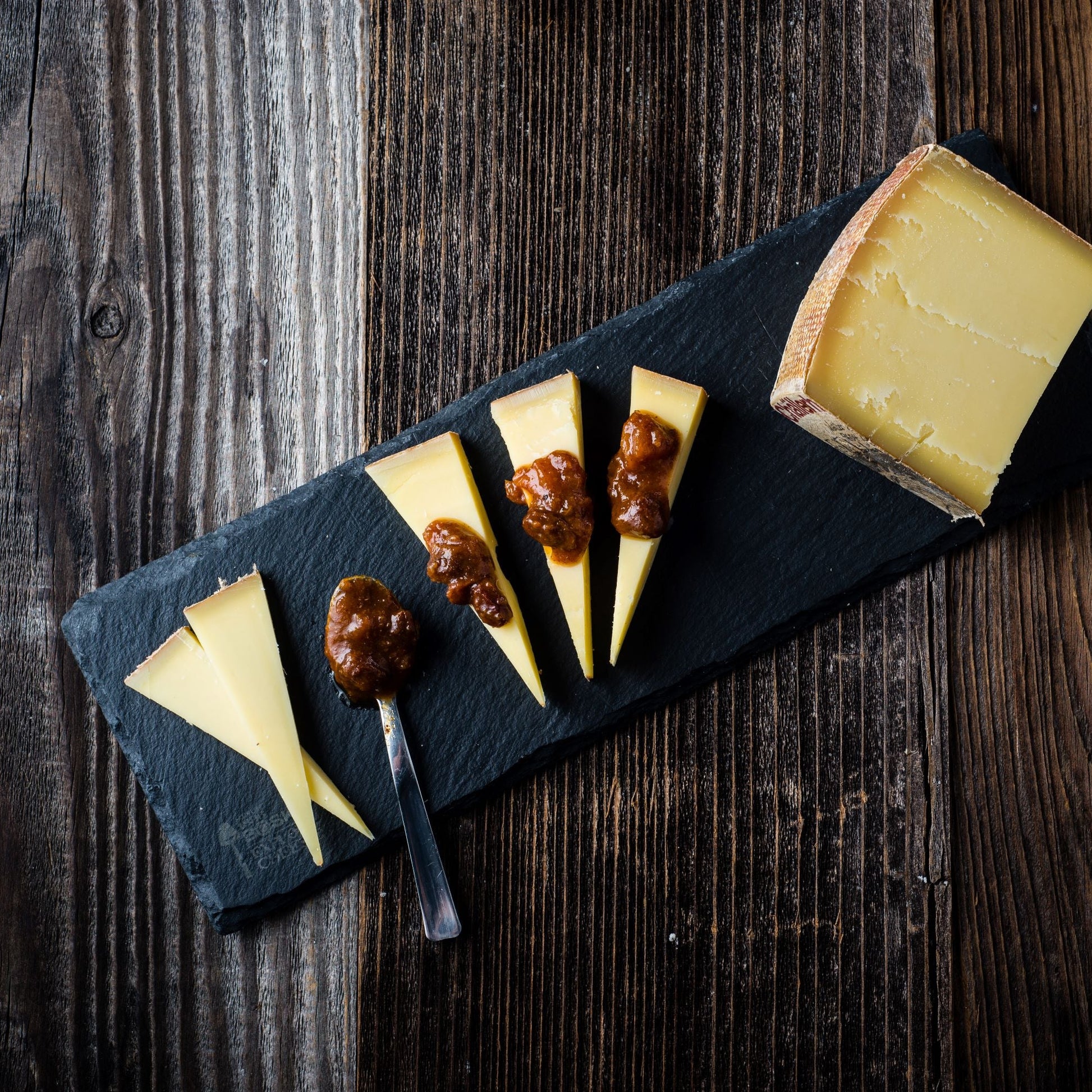 Cheese platter with sliced cheese and condiments on a dark slate board over a wooden background