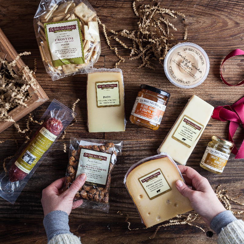 Assorted Di Bruno Bros. cheese and charcuterie items on a wooden surface with hands holding a piece of cheese.