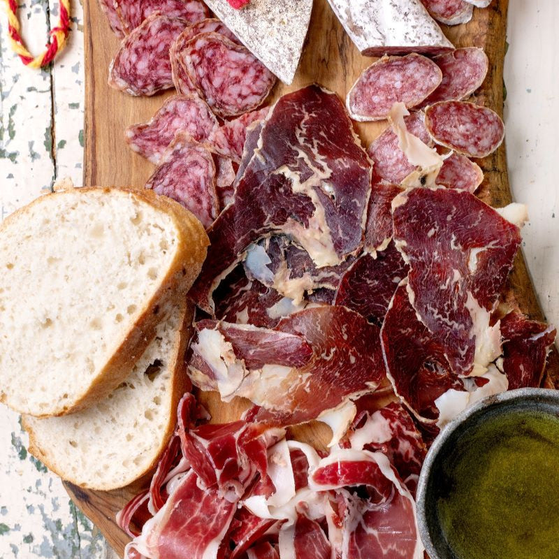 Platter of cured meats, bread, and nuts on a wooden board with a white background.