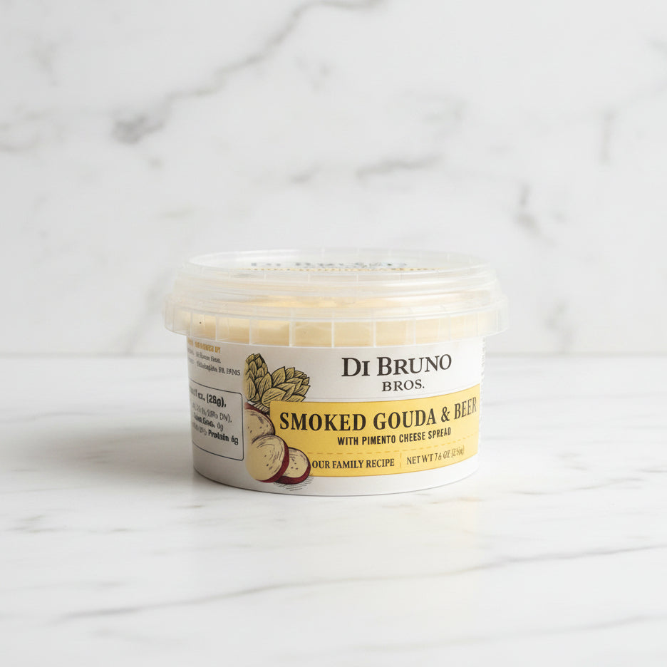 Container of Di Bruno Bros Smoked Gouda & Red Pepper Cheese on a dark surface with a tiled wall background.
