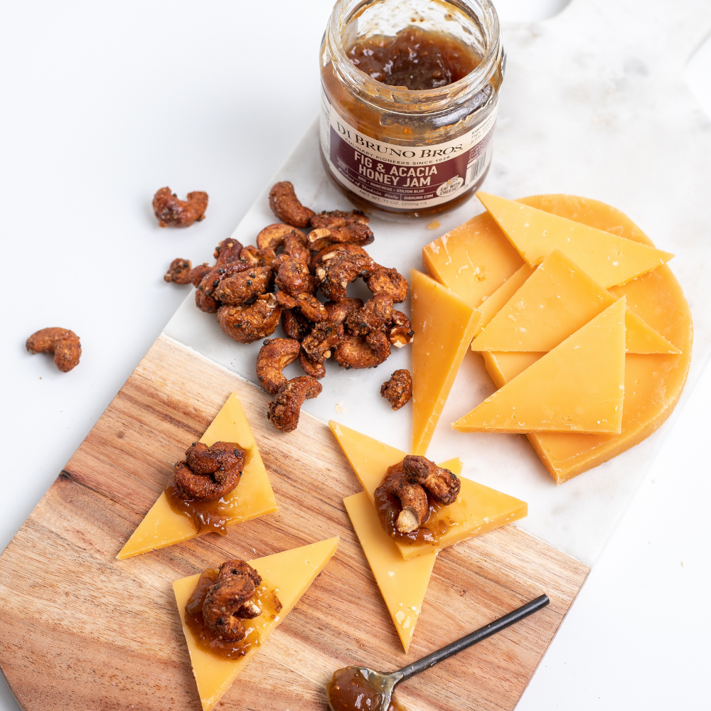Wooden board with cheese triangles topped with candied nuts and a jar of honey jam on a white background
