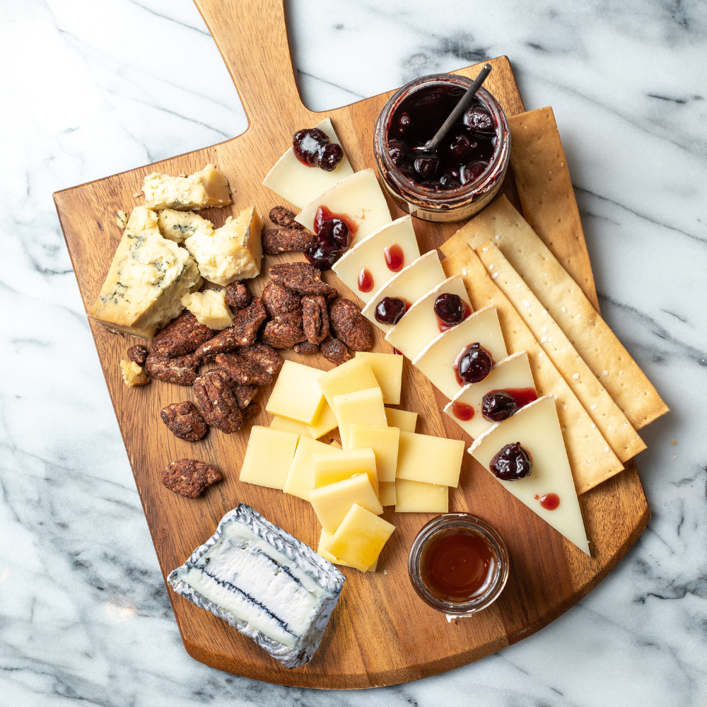 Wooden cheeseboard with various types of cheese, crackers, and accompaniments on a marble surface.