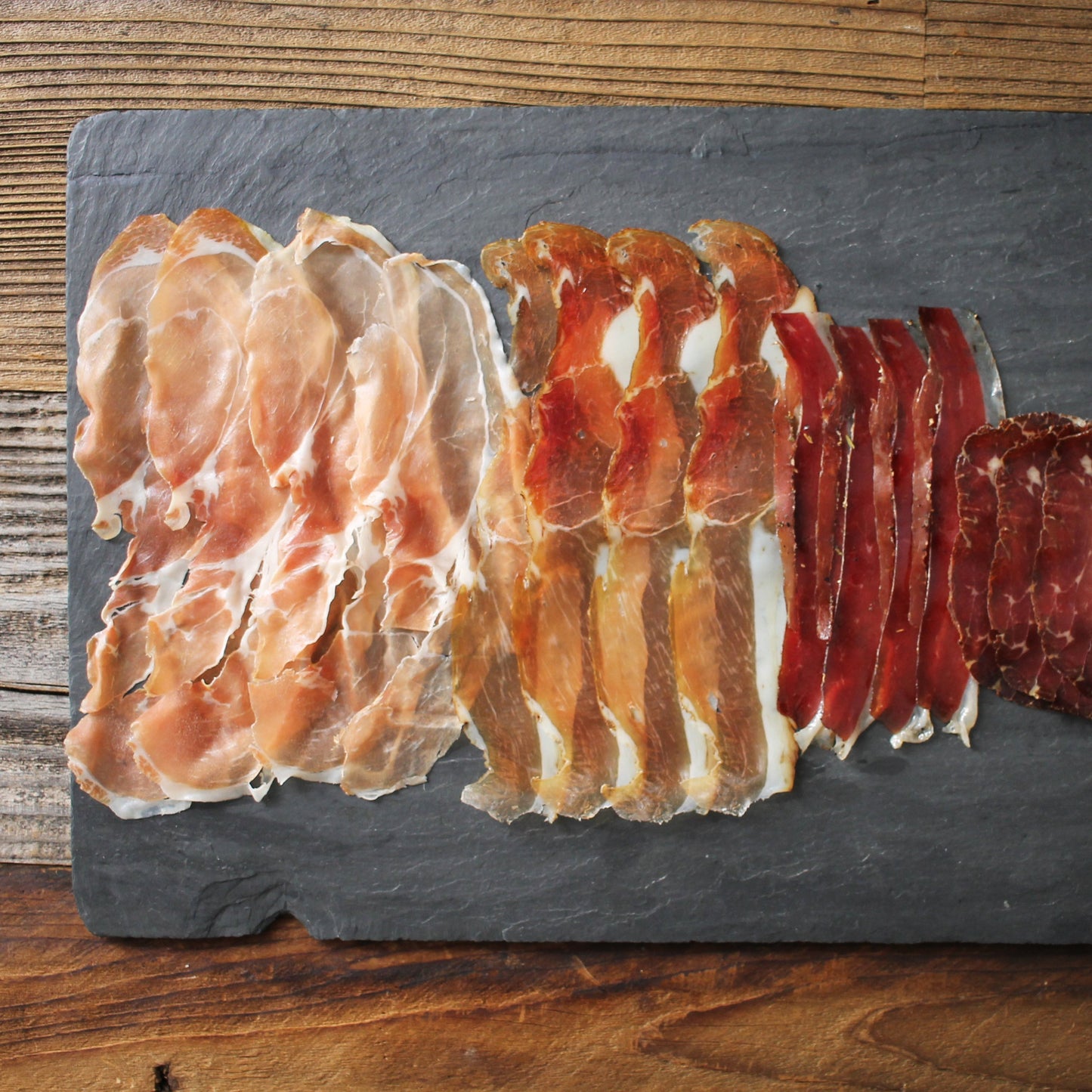 Three types of cured meats on a slate board over a wooden background