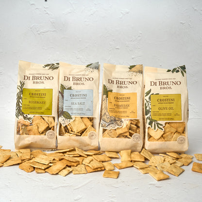 Four packages of Di Bruno Bros. Crostini Sampler Set with an assortment of flavors including olive oil, parmesan, rosemary, and sea salt, displayed in front of a white background with scattered crostini crackers.