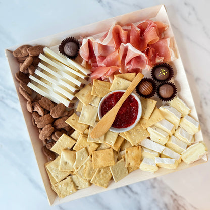 Assorted charcuterie board with meats, cheeses, crackers, and chocolates on a marble surface.