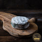 A wheel of Harbison cheese with a white rind, partially unwrapped from its spruce bark casing, sitting on a wooden board with a cheese knife.