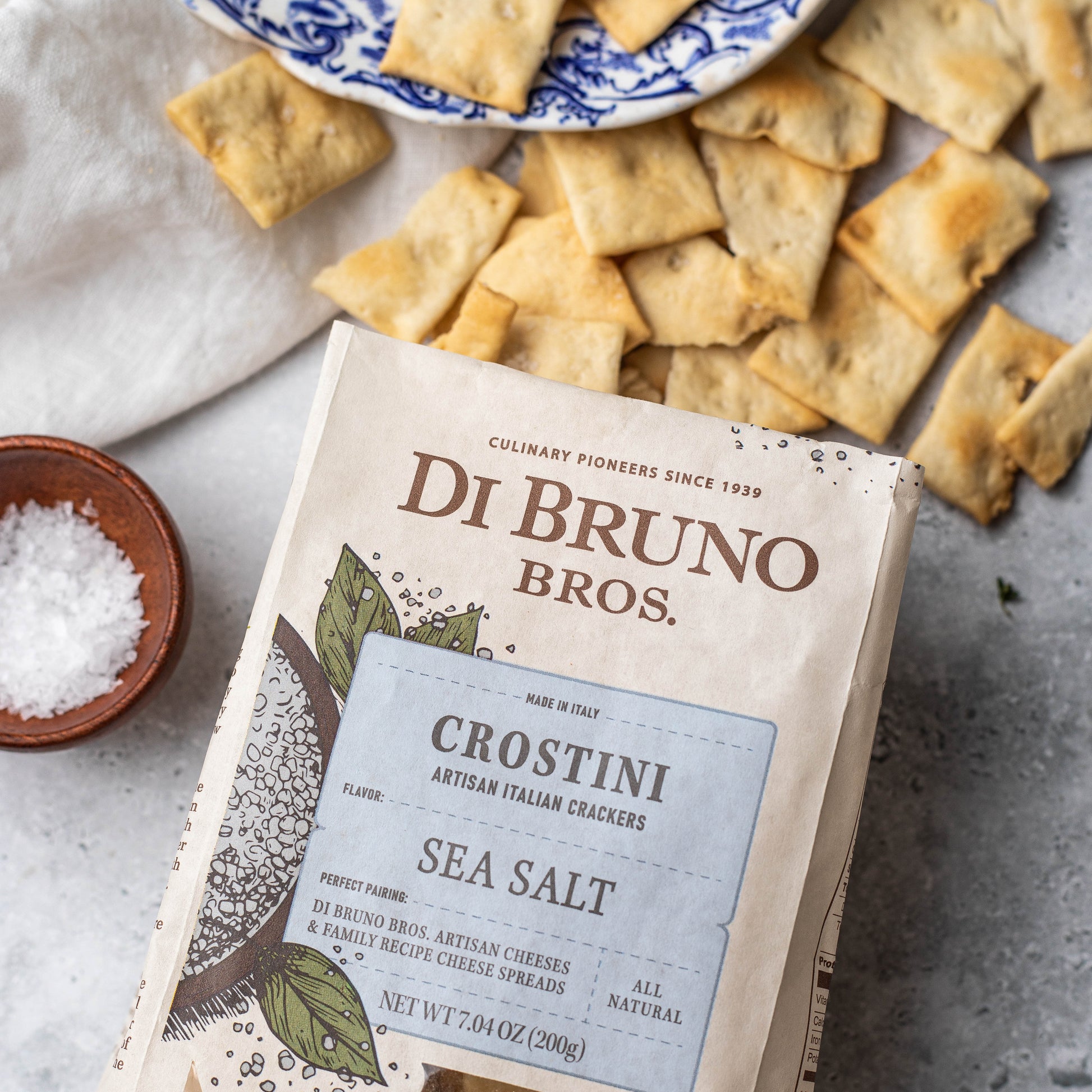 Di Bruno Bros. Crostini Sea Salt crackers packaging with crackers and a bowl of salt on a surface.