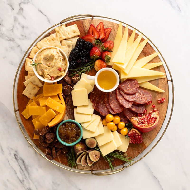 Styled round cheese and charcuterie board with sliced cheeses, salami, crostini, spreads, honey, fruit, and nuts