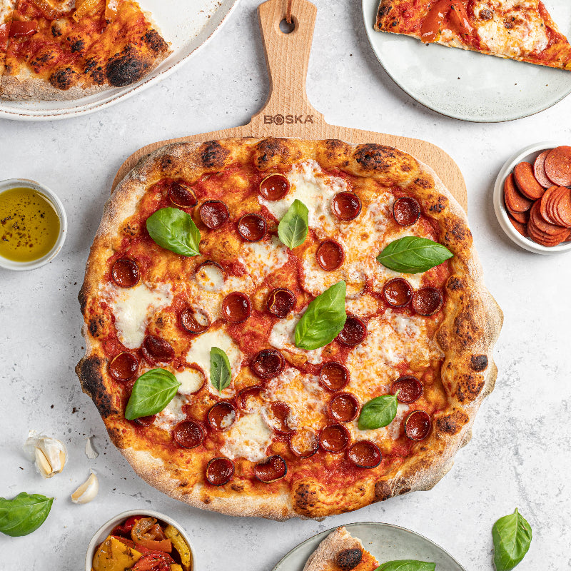 Wooden pizza peel with a pepperoni pizza on a light gray surface, surrounded by slices of pizza and ingredients.