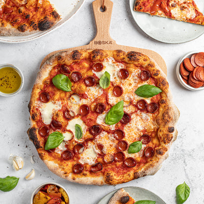 Wooden pizza peel with a pepperoni pizza on a light gray surface, surrounded by slices of pizza and ingredients.