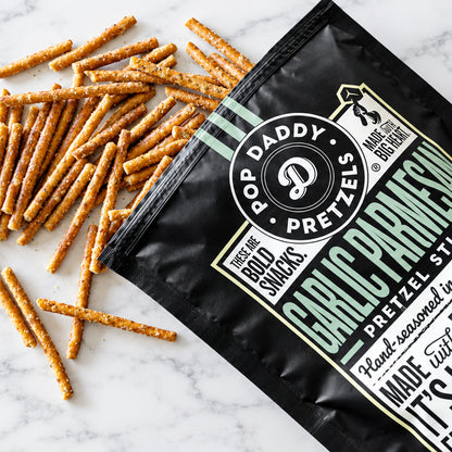 A bag of Pop Daddy Pretzels Garlic Parmesan flavor with pretzels spilled out on a white wood background.